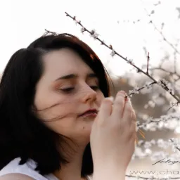 Portret młodej kobiety z ciemnymi włosami, delikatnie dotykającej kwitnącej gałązki drzewa, z widocznym logo fotografa w prawym dolnym rogu.