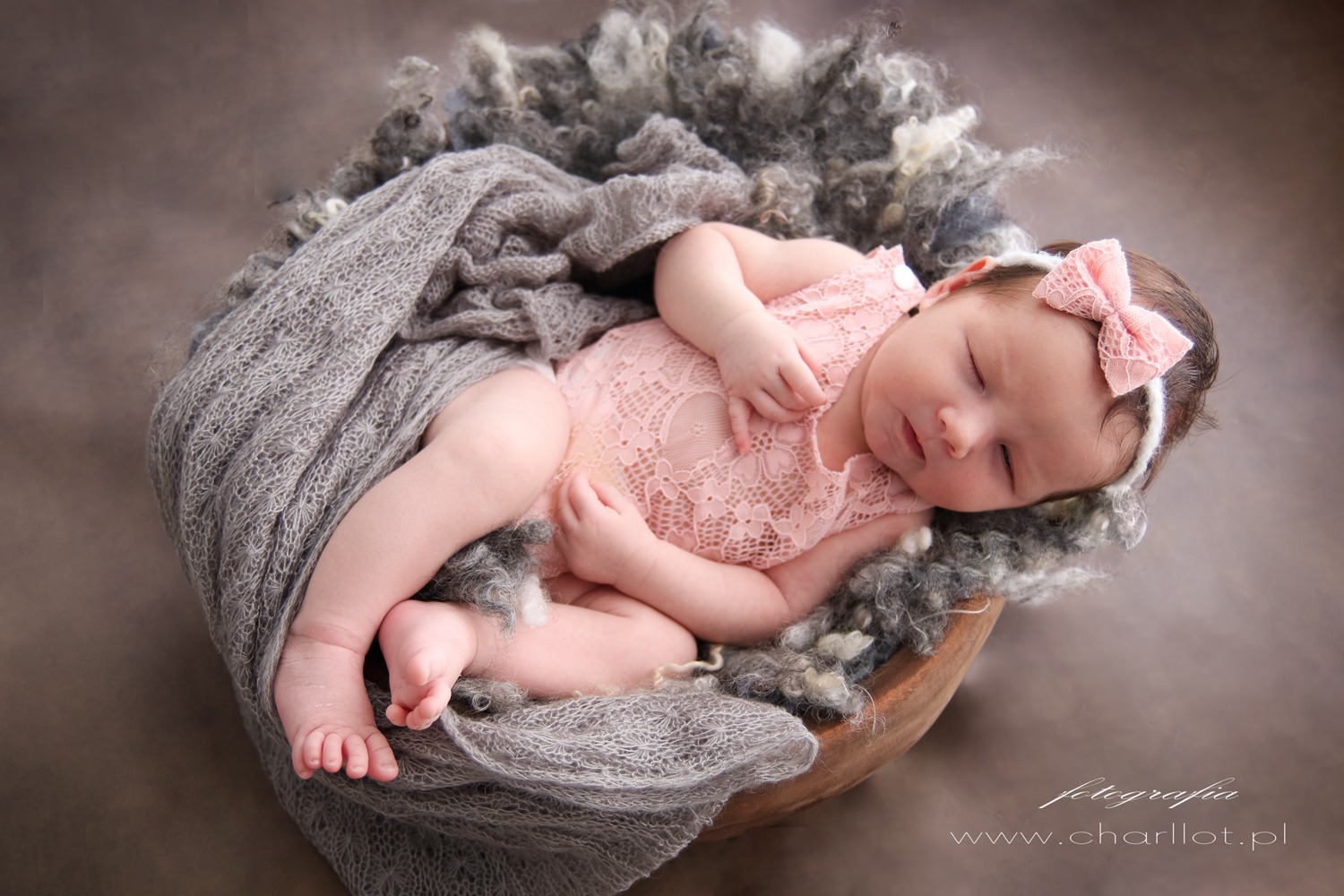 Śpiąca noworodek w różowym koronkowym body i opasce z kokardką, ułożona w drewnianej misie wyłożonej szarą wełną i dzierganym pledem.