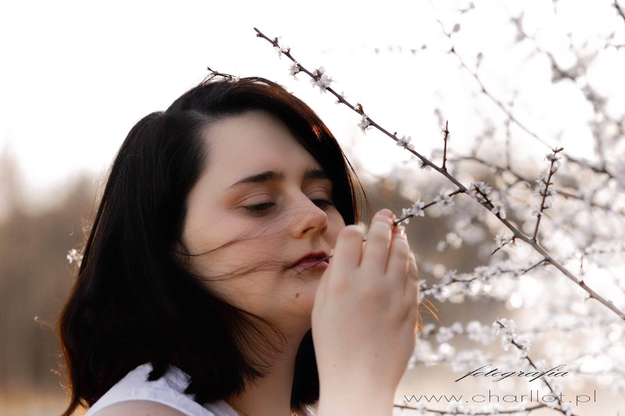 Portret młodej kobiety z ciemnymi włosami, delikatnie dotykającej kwitnącej gałązki drzewa, z widocznym logo fotografa w prawym dolnym rogu.