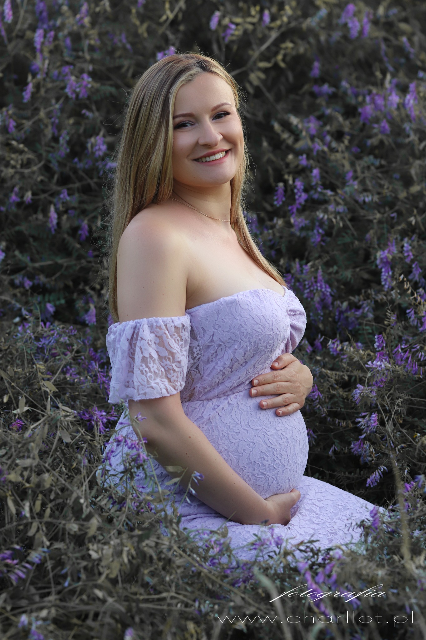 Uśmiechnięta, ciężarna blondynka w liliowej koronkowej sukience, siedząca w fioletowych kwiatach, delikatnie obejmująca dłońmi ciążowy brzuszek.