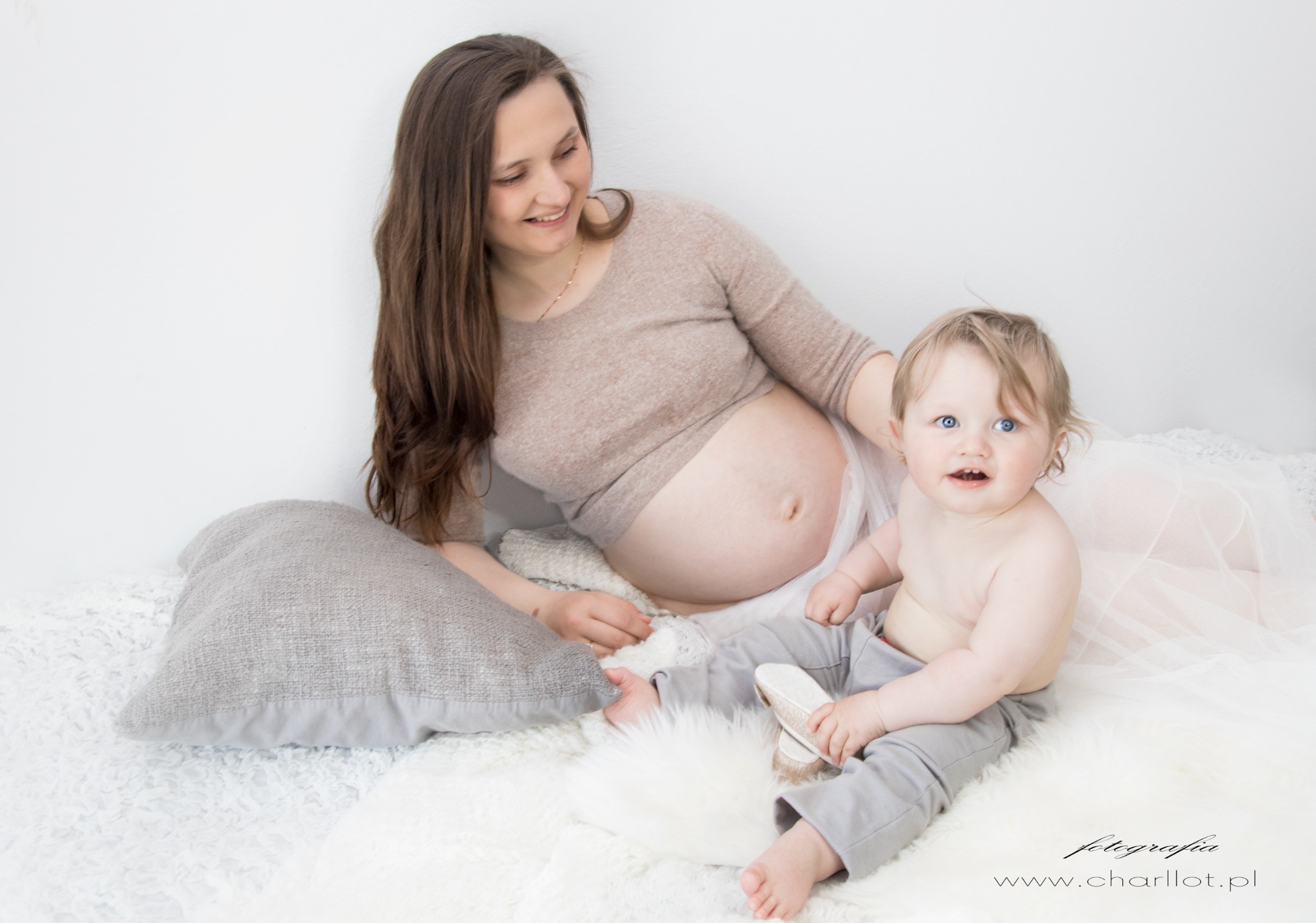 Uśmiechnięta, ciężarna kobieta w beżowym swetrze i tiulowej spódnicy leży na białym futrze obok małego dziecka w szarych spodniach, które trzyma mały, błyszczący but.
