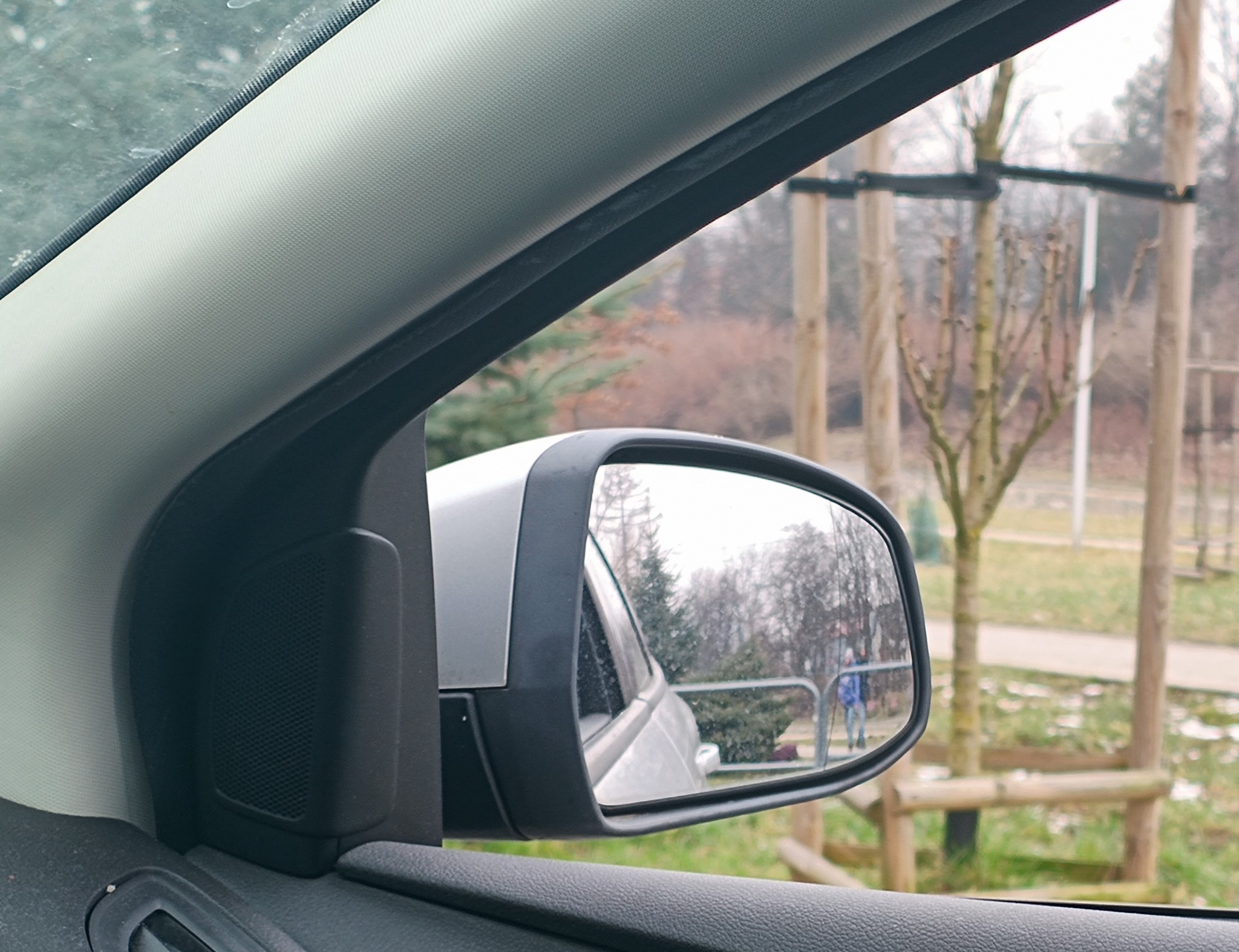 Widok z wnętrza samochodu na lusterko boczne, w którym odbija się fragment ulicy z drzewami i przechodniem w niebieskiej kurtce.