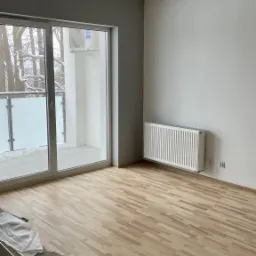 Jasne panele ułożone w pustym pokoju z grzejnikiem i balkonem, widoczne ślady prac wykończeniowych przy listwie przypodłogowej.