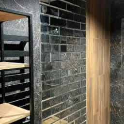 Ściana wyłożona lustrzanymi, prostokątnymi płytkami imitującymi cegłę, sąsiadująca z panelami drewnopodobnymi, na tle czarnego marmuru z złotymi żyłami oraz fragment czarnej metalowej konstrukcji...