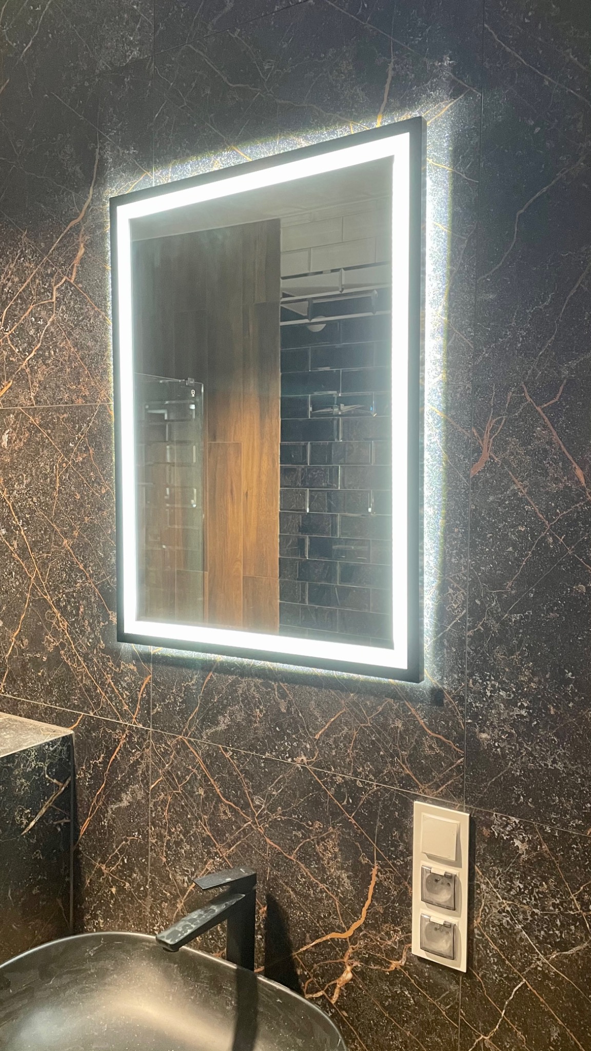 Nowoczesna łazienka z czarnymi płytkami imitującymi marmur, podświetlane lustro kwadratowe, czarna bateria umywalkowa i gniazdka elektryczne z klapkami.