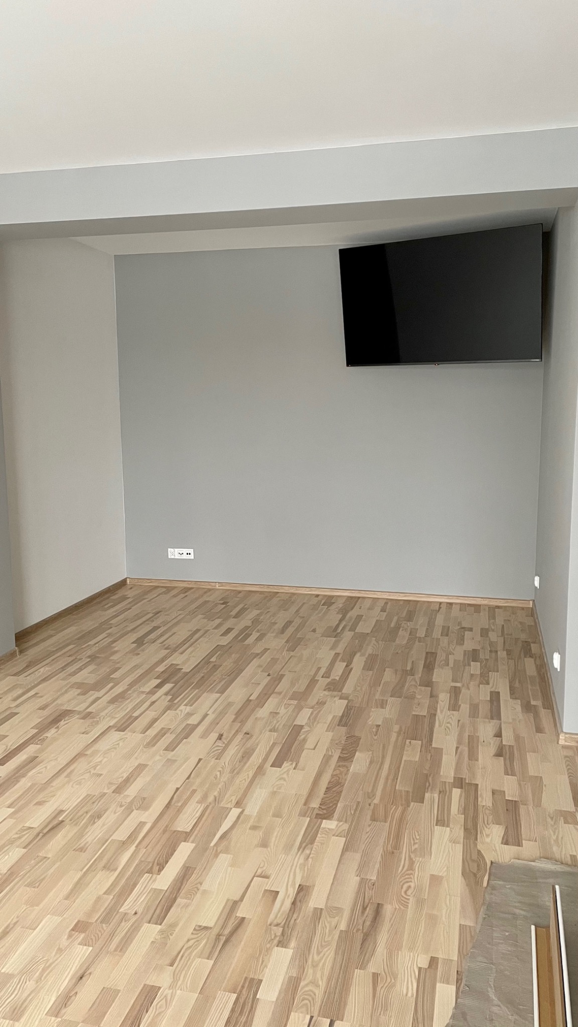 Jasny, świeżo położony parkiet w pustym pokoju z szarymi ścianami i wiszącym telewizorem, widoczne listwy przypodłogowe i gniazdka elektryczne.
