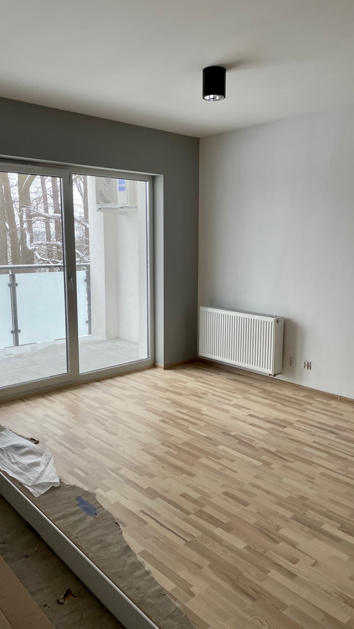 Jasne panele ułożone w pustym pokoju z grzejnikiem i balkonem, widoczne ślady prac wykończeniowych przy listwie przypodłogowej.