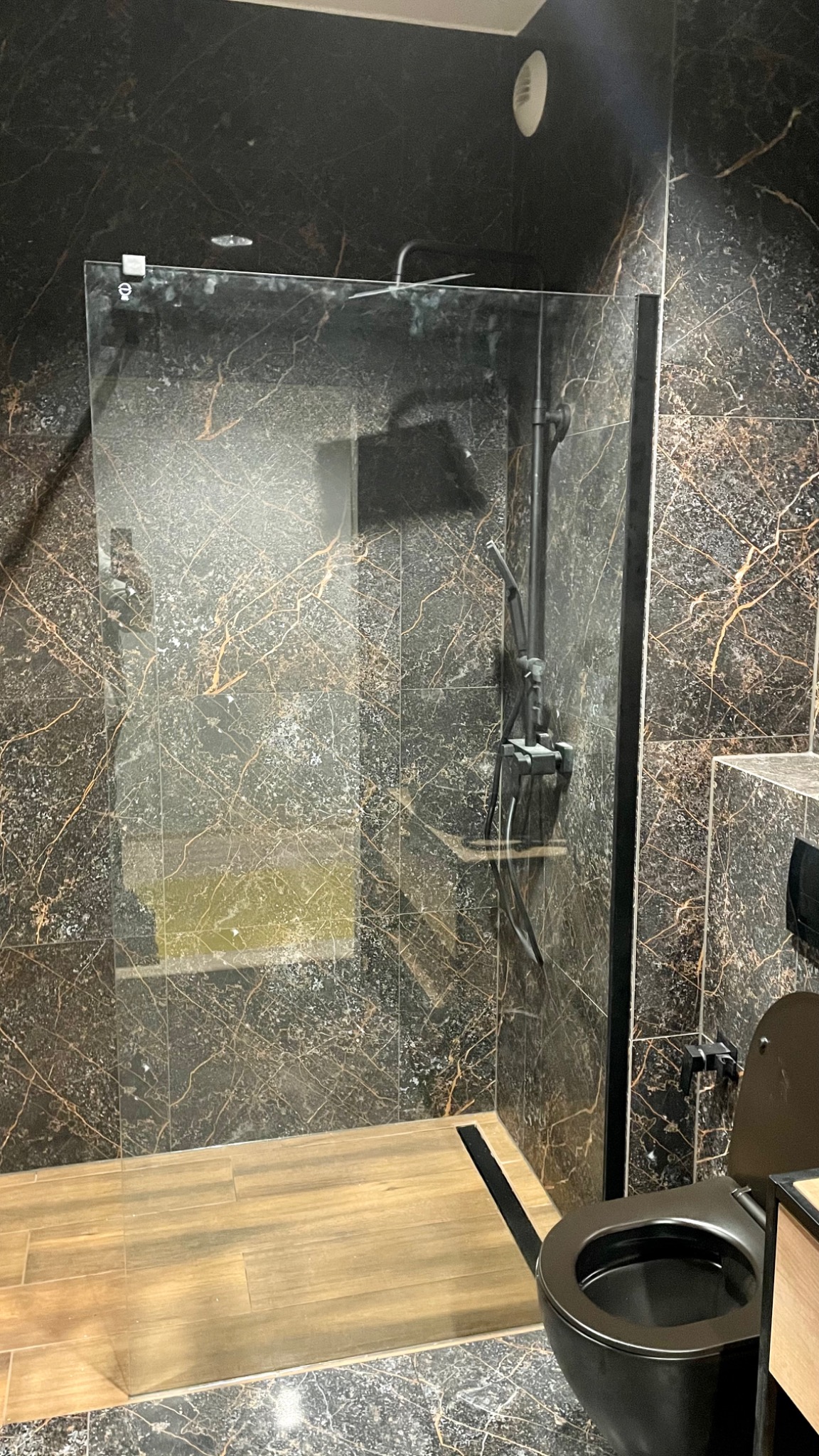 Nowoczesna łazienka z kabiną prysznicową ze szklaną ścianką, czarnymi płytkami imitującymi marmur ze złotymi żyłami, drewnopodobną podłogą i czarną armaturą.