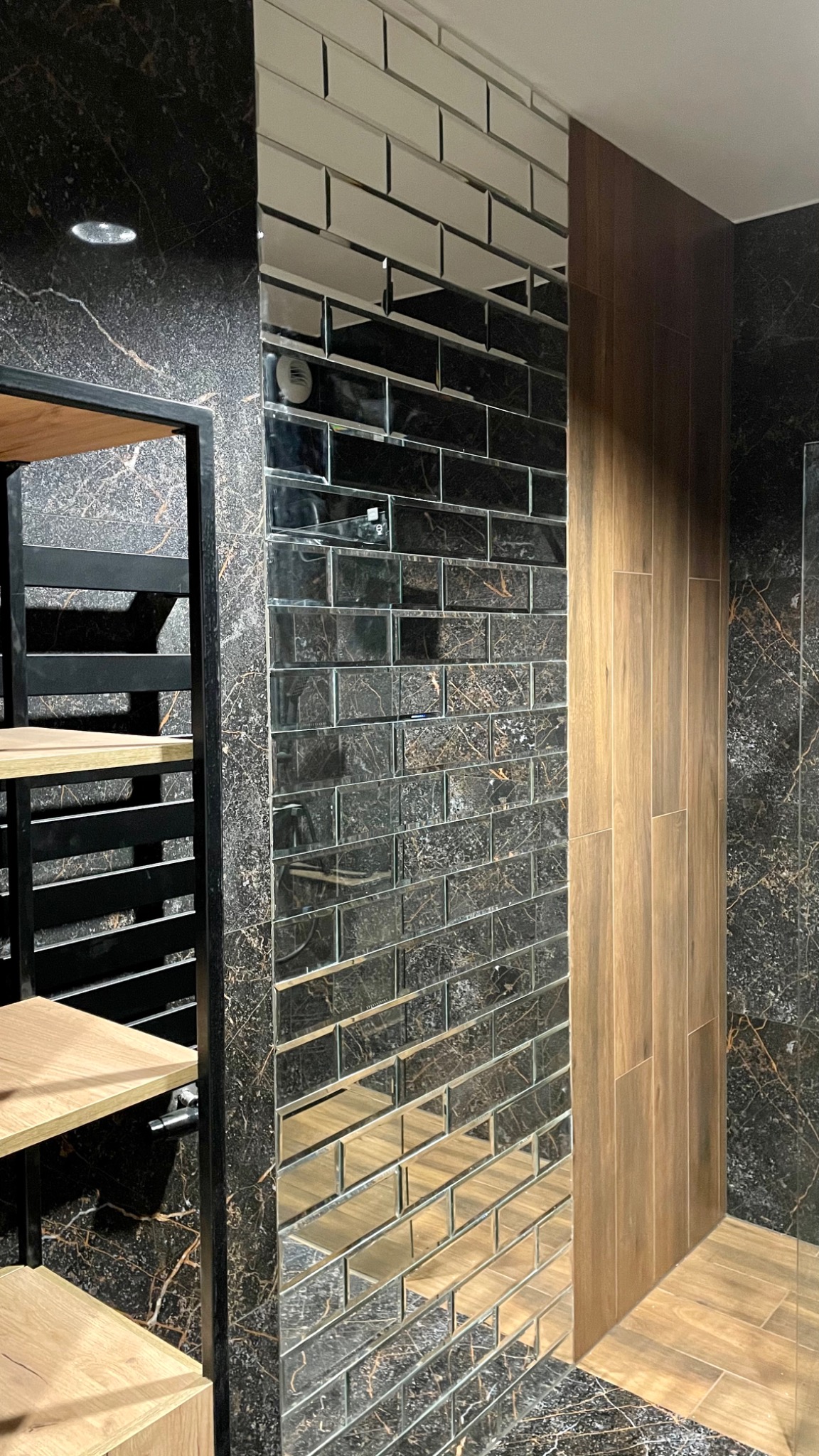 Ściana wyłożona lustrzanymi, prostokątnymi płytkami imitującymi cegłę, sąsiadująca z panelami drewnopodobnymi, na tle czarnego marmuru z złotymi żyłami oraz fragment czarnej metalowej konstrukcji...