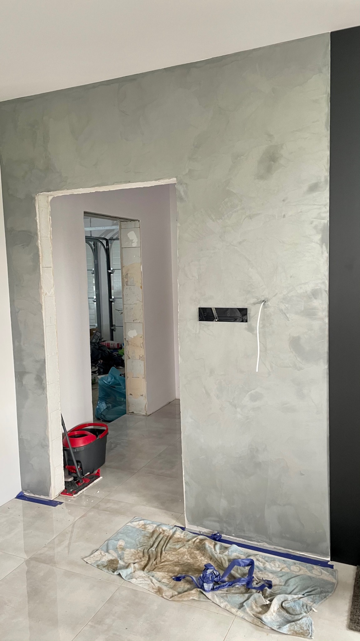 Świeżo pomalowana ściana z efektem dekoracyjnym w odcieniach szarości, widoczny włącznik elektryczny z wystającym przewodem, na podłodze leży brudny ręcznik i niebieska folia zabezpieczająca, w tle...