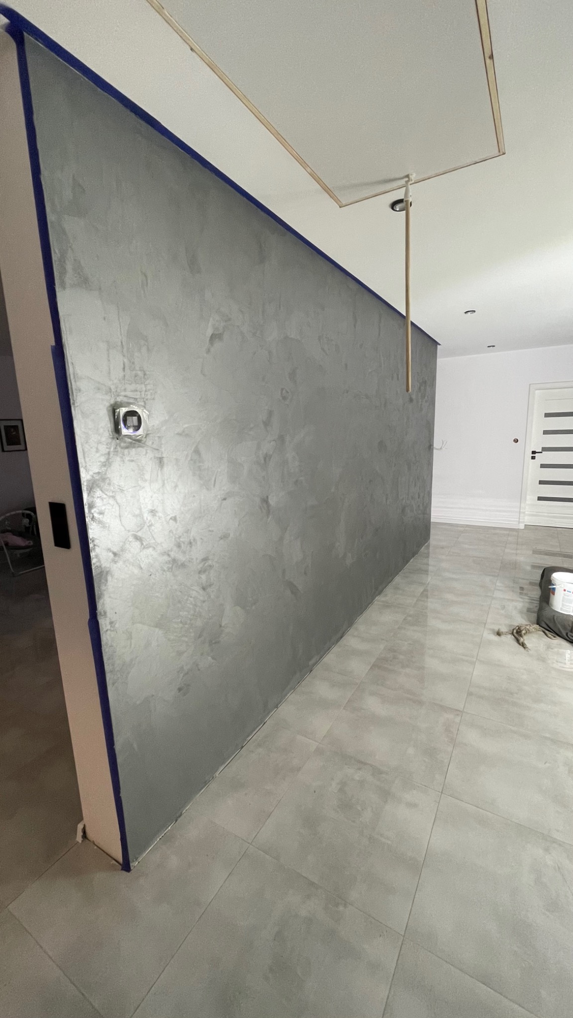 Ściana z efektem dekoracyjnym w odcieniach szarości, zabezpieczona taśmą malarską, z widocznym gniazdkiem elektrycznym i fragmentem sufitu z otworem rewizyjnym.