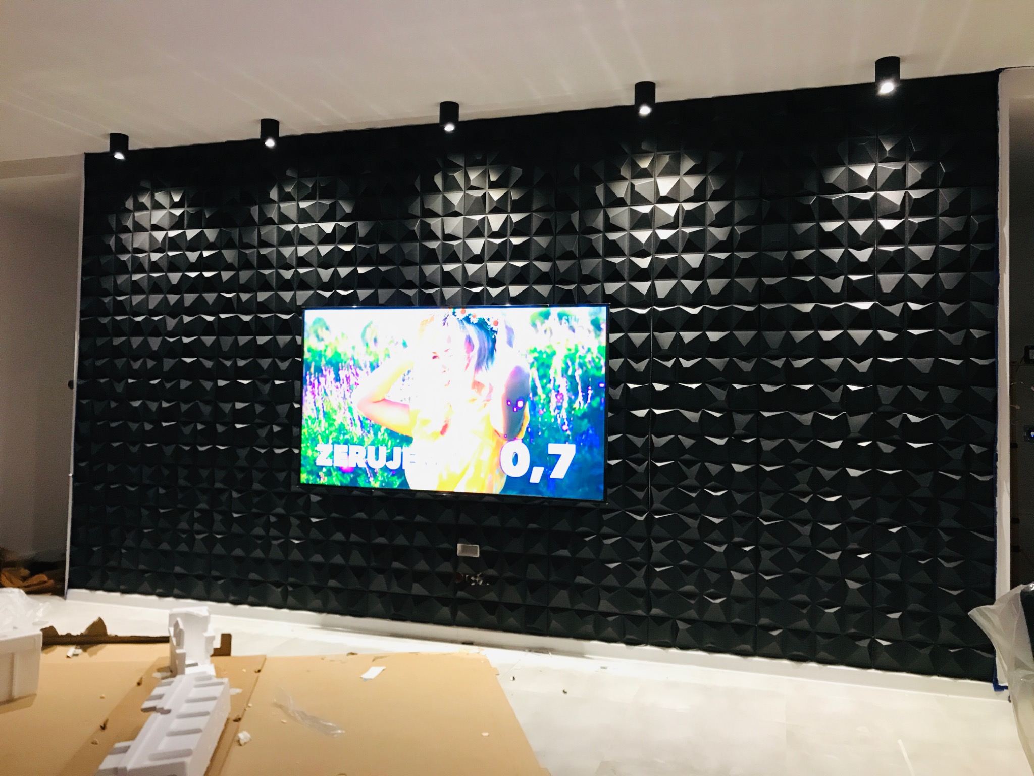 Czarna ściana z trójwymiarowych paneli dekoracyjnych, oświetlona sześcioma punktowymi lampami sufitowymi, z zawieszonym telewizorem wyświetlającym kolorowy obraz; na podłodze kartony i pozostałości...