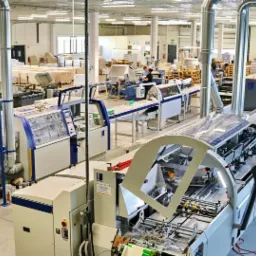 Wnętrze hali drukarni z linią produkcyjną do druku i obróbki papieru, widoczne palety z papierem i pracownicy obsługujący maszyny.