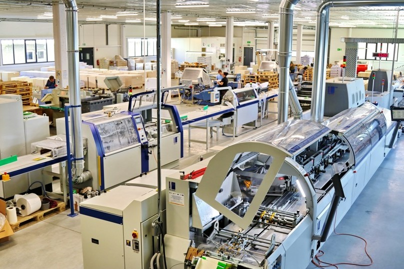 Wnętrze hali drukarni z linią produkcyjną do druku i obróbki papieru, widoczne palety z papierem i pracownicy obsługujący maszyny.
