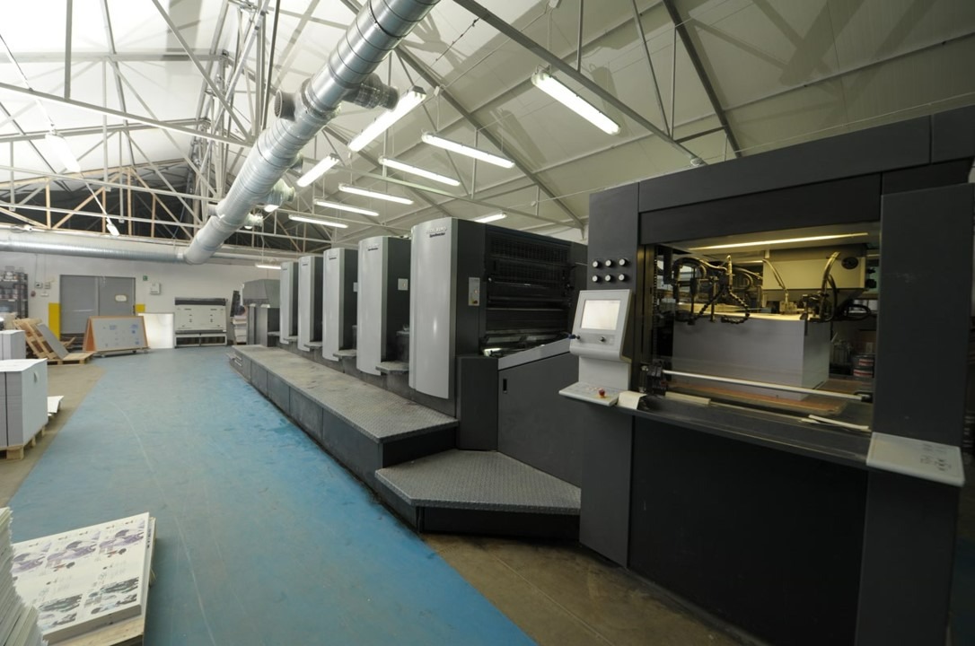 Wnętrze drukarni z dużą, wielokolorową maszyną offsetową Heidelberg Speedmaster w trakcie pracy, widoczne stosy papieru i system wentylacji pod sufitem.
