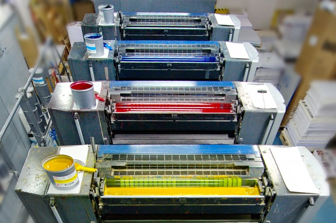 Maszyna offsetowa w trakcie pracy, z widocznymi pojemnikami z farbami: żółtą, czerwoną, niebieską i czarną.