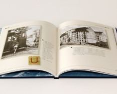 Otwarta książka z fotografiami i tekstem, leżąca na białym tle. Widoczne zdjęcia przedstawiają budynki i pojazdy, a na jednej ze stron umieszczono złoty element graficzny w kształcie litery U.