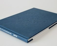 Ciemnoniebieska książka w twardej oprawie z wytłoczonym wzorem na okładce, leżąca na białym tle. Widoczny grzbiet z napisem.