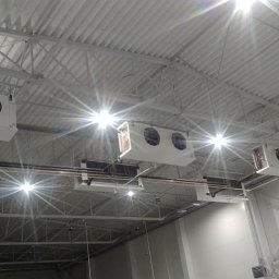 ART COOL Adam Czerwński - Białe chłodnice sufitowe z wentylatorami w hali przemysłowej, widoczne rury i oświetlenie, perspektywa z dołu.