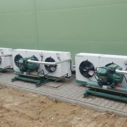 ART COOL Adam Czerwński - Trzy agregaty chłodnicze na zewnątrz budynku z zieloną elewacją, ustawione na szarej kostce brukowej. Widoczne wentylatory i rury chłodnicze.