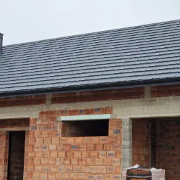 Nowy, grafitowy dach z dachówki ceramicznej na budynku w stanie surowym otwartym, widoczny komin i orynnowanie.