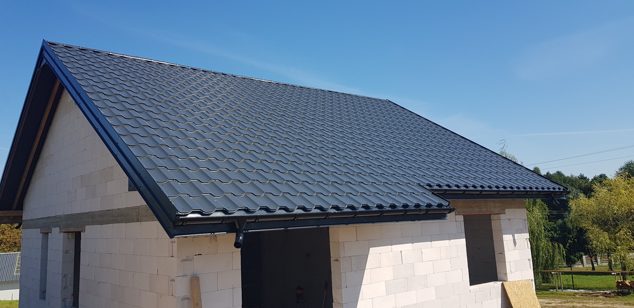 Nowe, ciemnoszare pokrycie dachowe z blachy na domu w trakcie budowy, widoczne białe bloczki i niezakończone otwory okienne, błękitne niebo w tle.