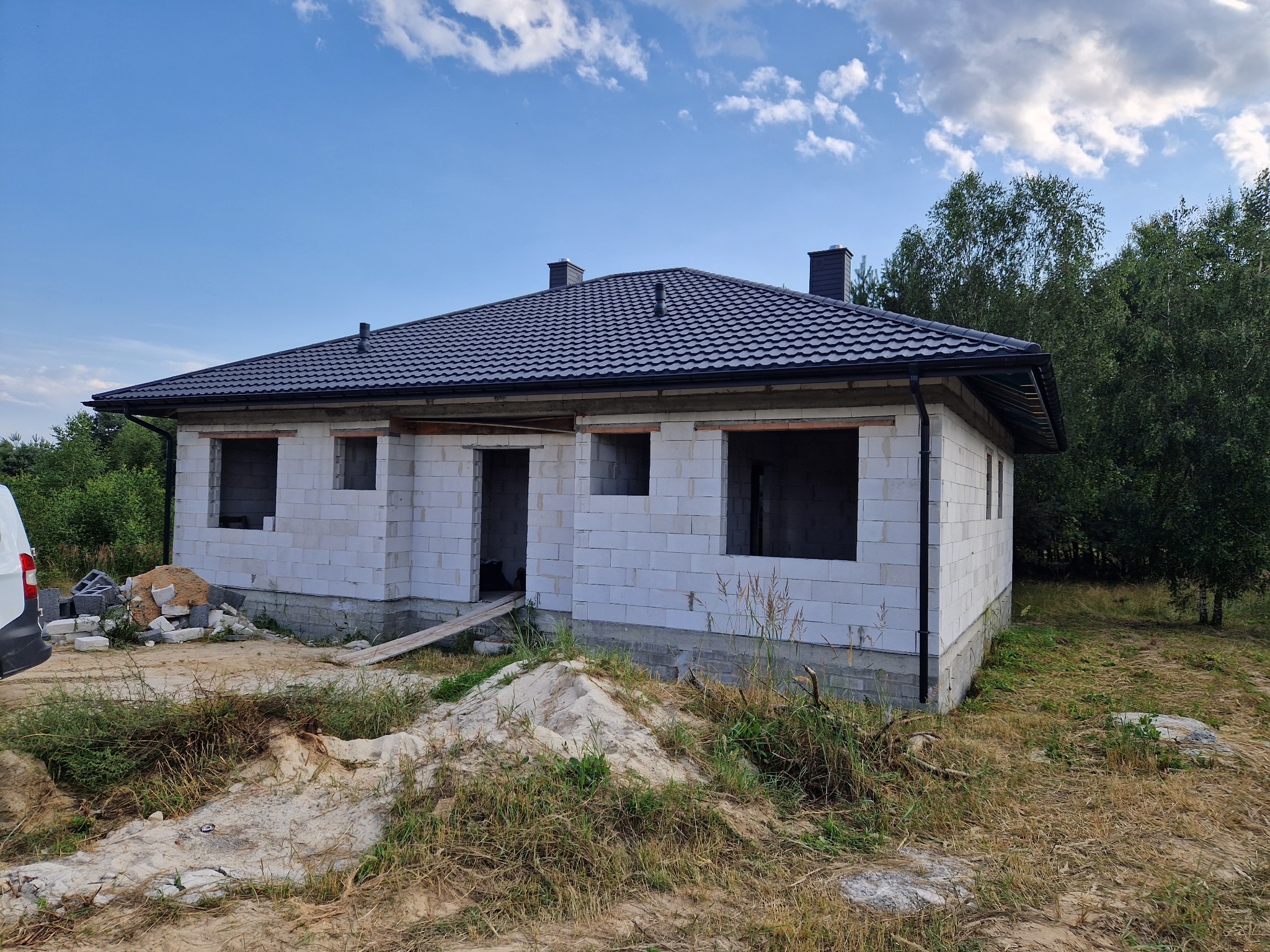 Dom w trakcie budowy z zamontowanym, nowym, czarnym dachem z dachówki modułowej, widoczne niezabezpieczone otwory okienne i drzwiowe, oraz pozostałości materiałów budowlanych.