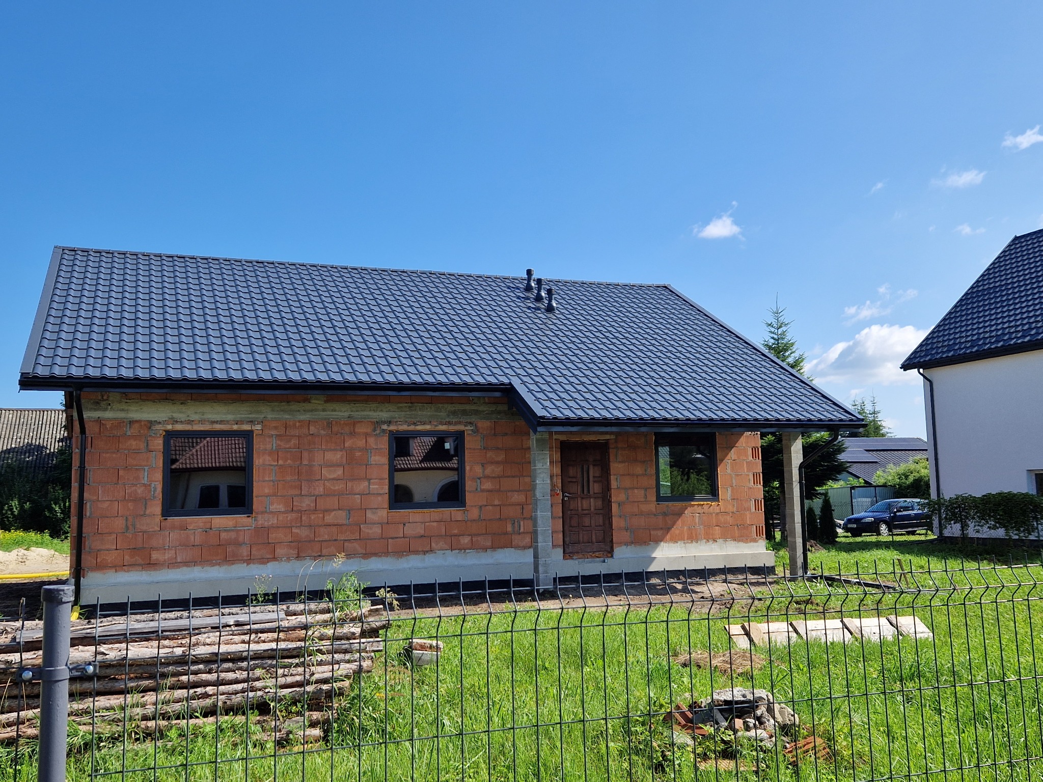 Niedokończony budynek jednorodzinny z ciemnoszarą, metalową konstrukcją pokrycia na dachu, widoczne kominy, ściany z czerwonej cegły, czarne ramy okienne, drewniane drzwi wejściowe, w tle drugi...