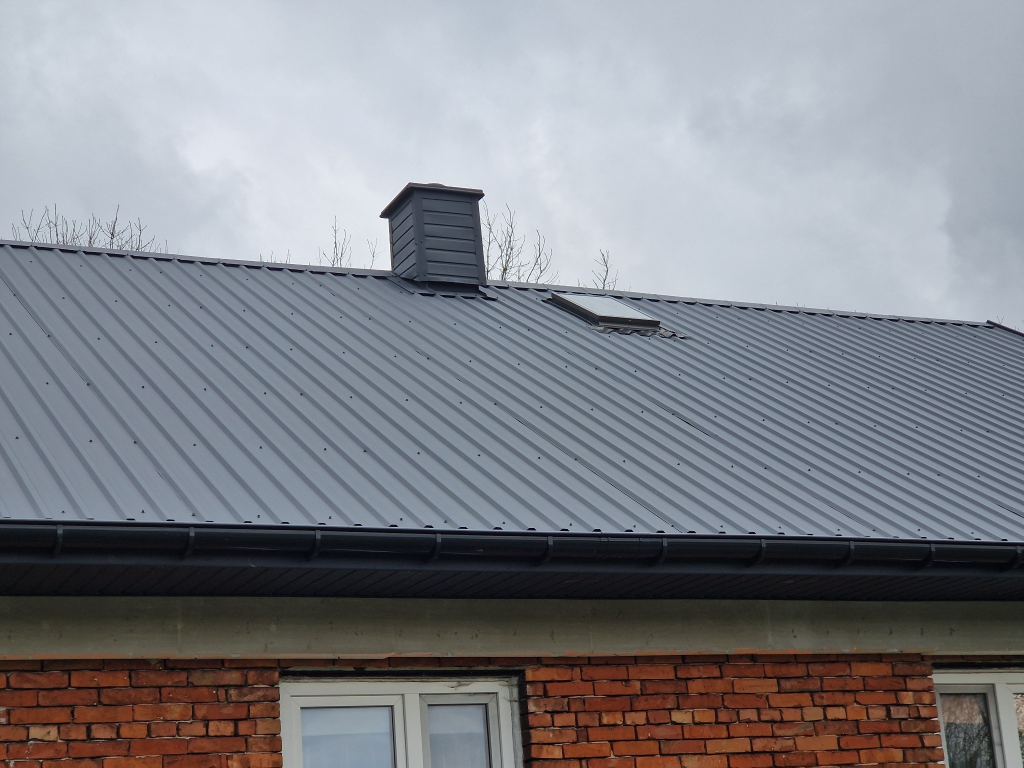 Szary dach z blachy, widoczny komin i okno dachowe, budynek z czerwonej cegły w pochmurny dzień.