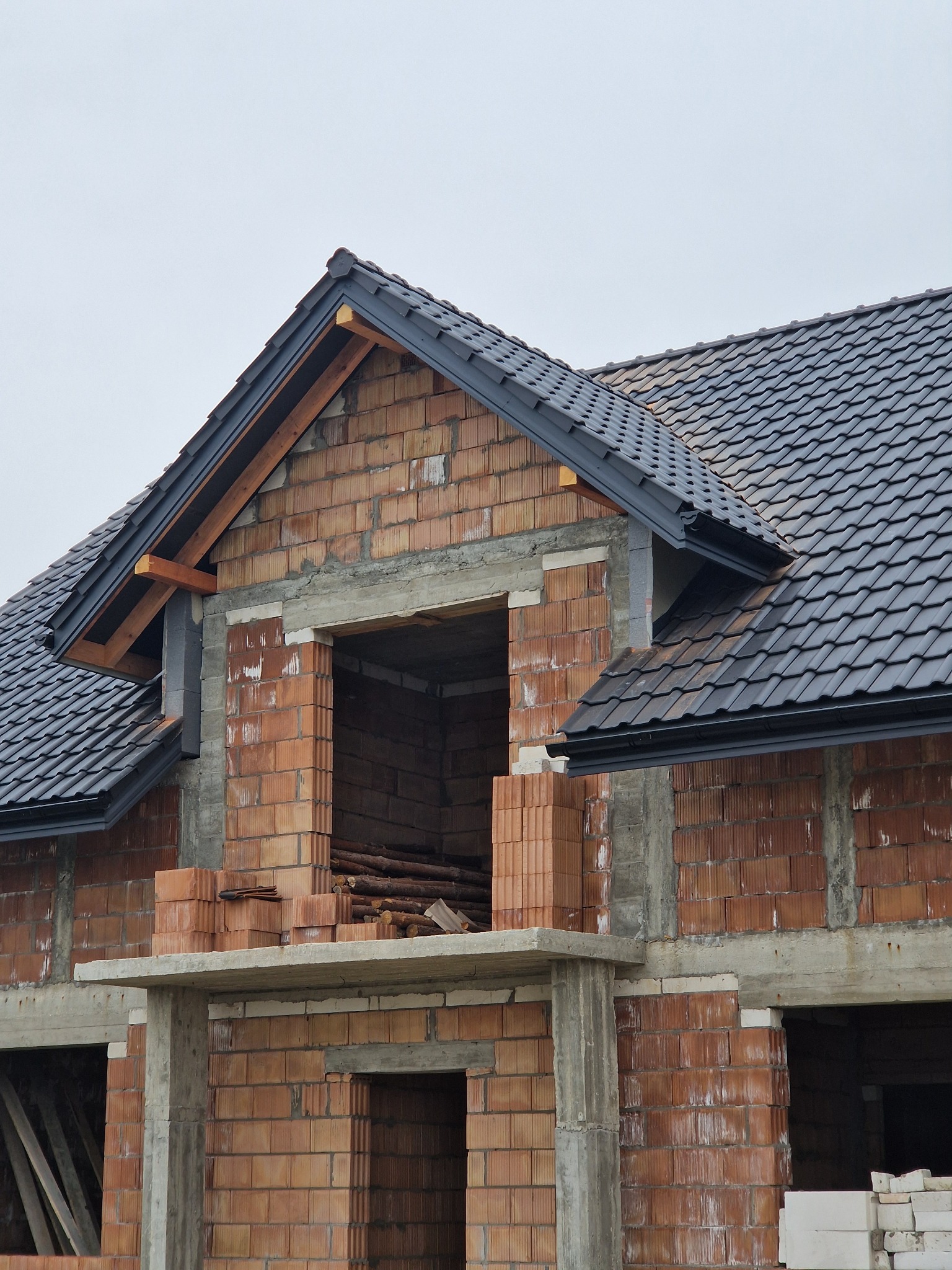 Budynek w trakcie budowy, z widoczną konstrukcją dachu pokrytą ciemną dachówką ceramiczną i fragmentami muru z cegły dziurawki.