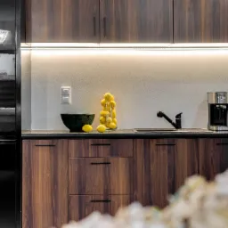 Nowoczesna kuchnia z ciemnymi, drewnianymi szafkami, czarnym blatem, czarną lodówką i stalowym ekspresem do kawy. Na blacie dekoracyjny zielony wazon i szklany słój wypełniony cytrynami.