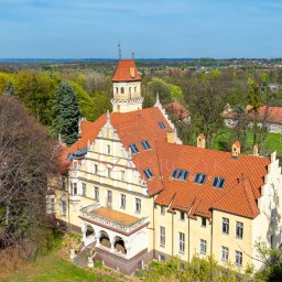 SZAFARCZYK NIERUCHOMOŚCI - Panorama z lotu ptaka na odrestaurowany, zabytkowy pałac z czerwoną dachówką, otoczony parkiem i lasem w słoneczny dzień.