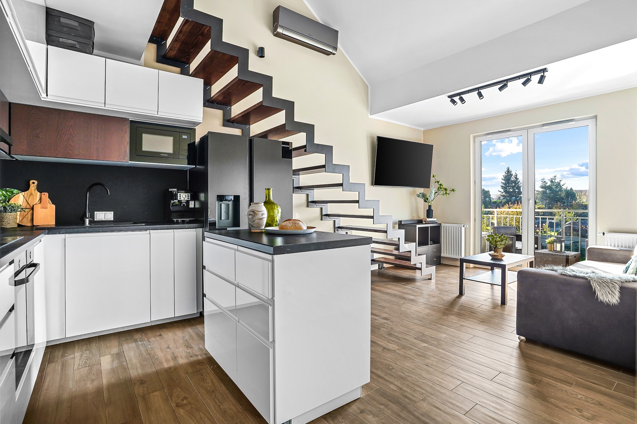 Nowoczesna kuchnia z wyspą i schodami ażurowymi, czarne blaty, białe fronty, drewniane podłogi, widok na salon z dużym oknem balkonowym.