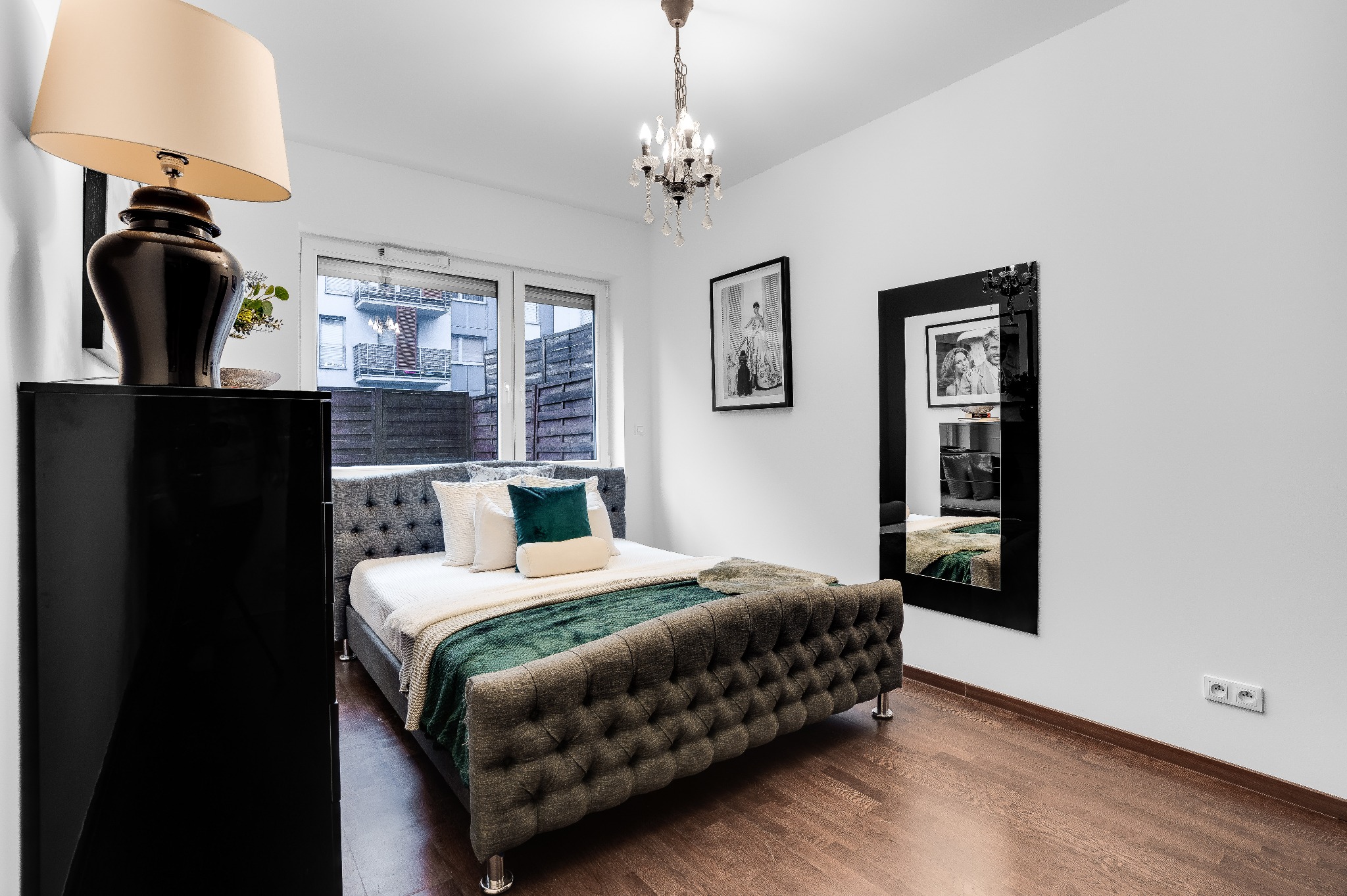 Elegancka sypialnia z szarym tapicerowanym łóżkiem, zielonym pledem i dekoracyjnymi poduszkami, oświetlona kryształowym żyrandolem i lampą stojącą na czarnej komodzie, z lustrem w czarnej ramie...