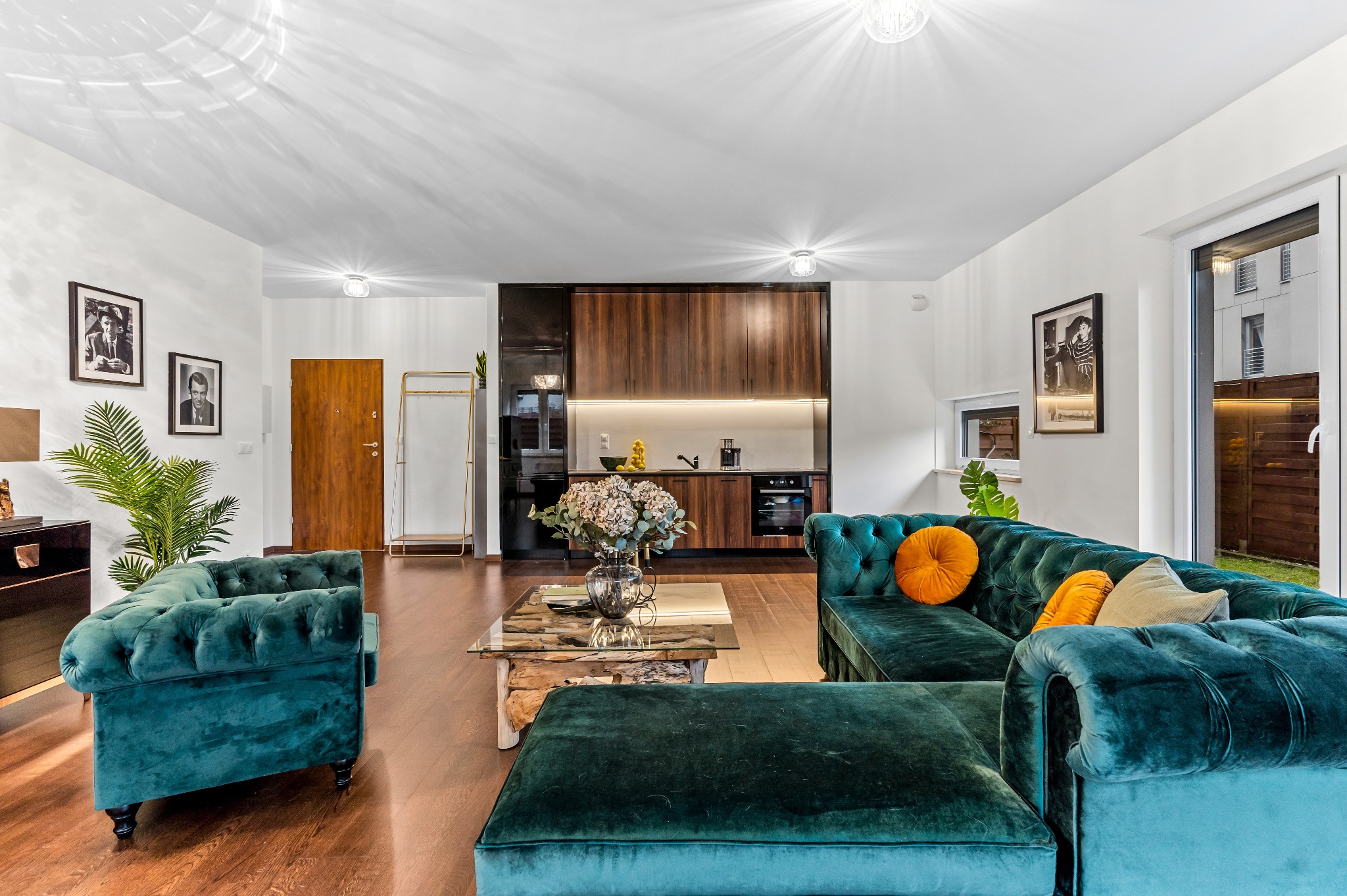 Salon z otwartą kuchnią, turkusowe sofy Chesterfield, szklany stolik kawowy z drewnianą podstawą, drewniane podłogi, czarno-brązowe szafki kuchenne z oświetleniem LED podszafkowym.