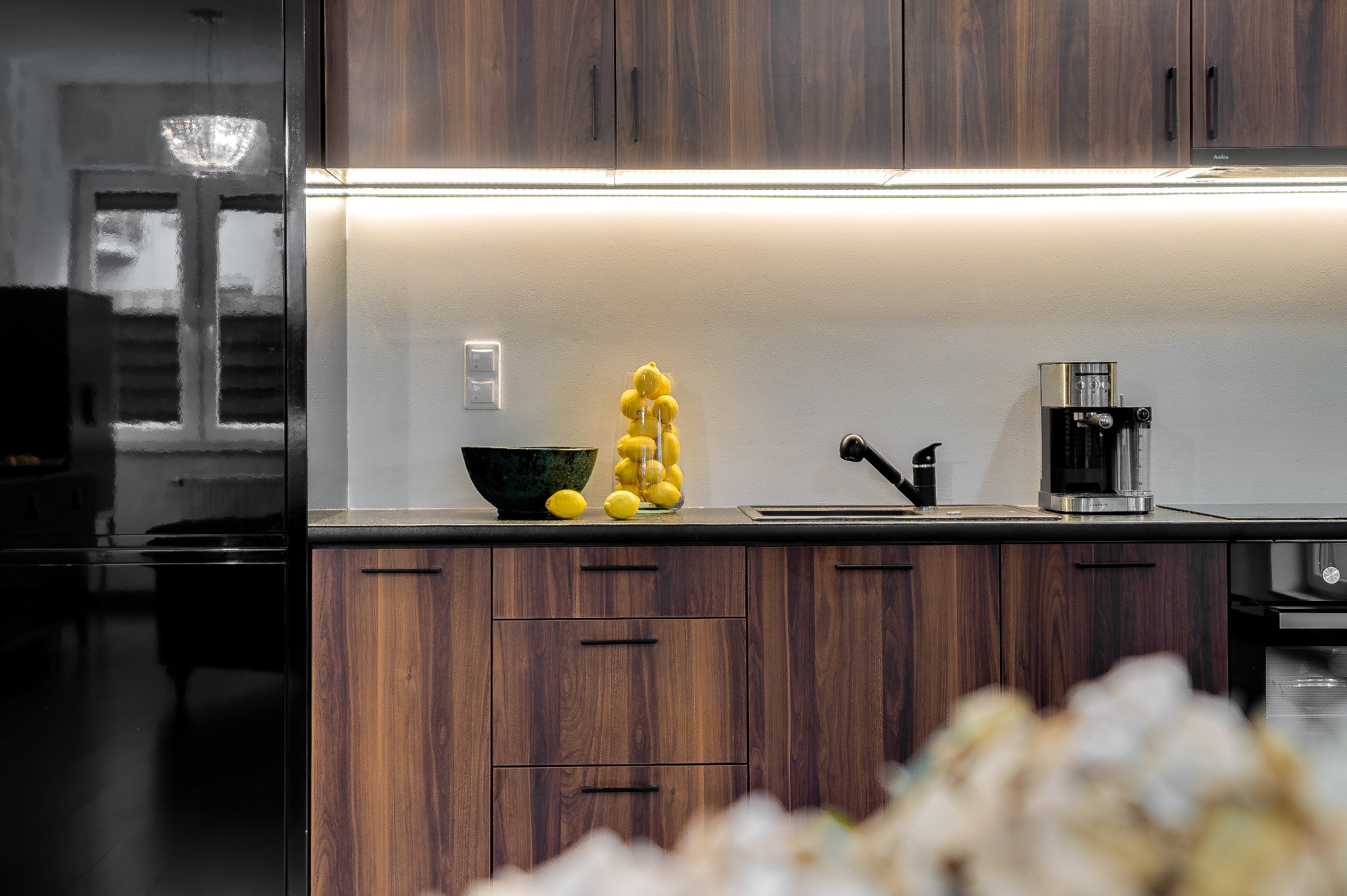 Nowoczesna kuchnia z ciemnymi, drewnianymi szafkami, czarnym blatem, czarną lodówką i stalowym ekspresem do kawy. Na blacie dekoracyjny zielony wazon i szklany słój wypełniony cytrynami.
