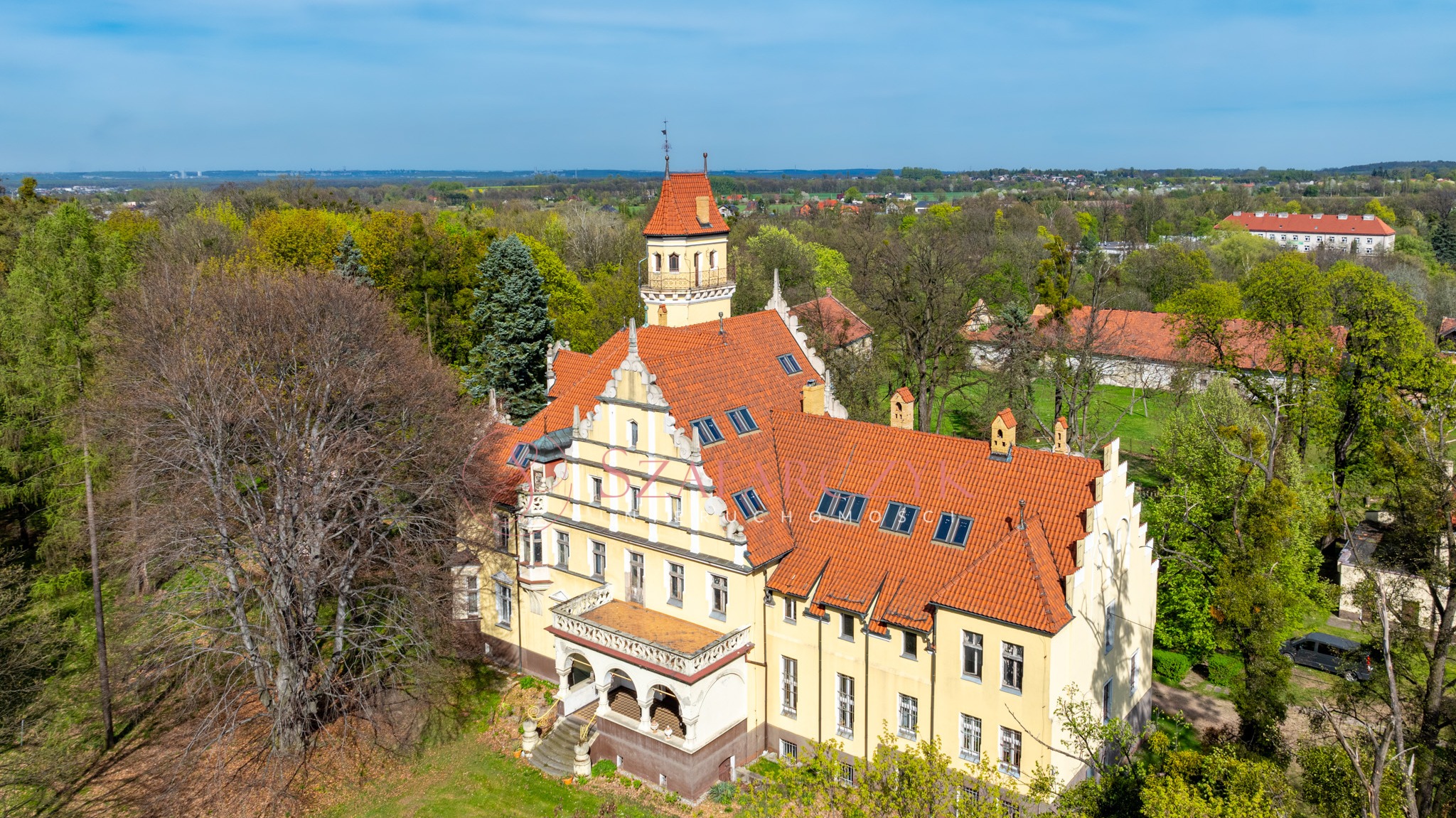 Panorama z lotu ptaka na odrestaurowany, zabytkowy pałac z czerwoną dachówką, otoczony parkiem i lasem w słoneczny dzień.