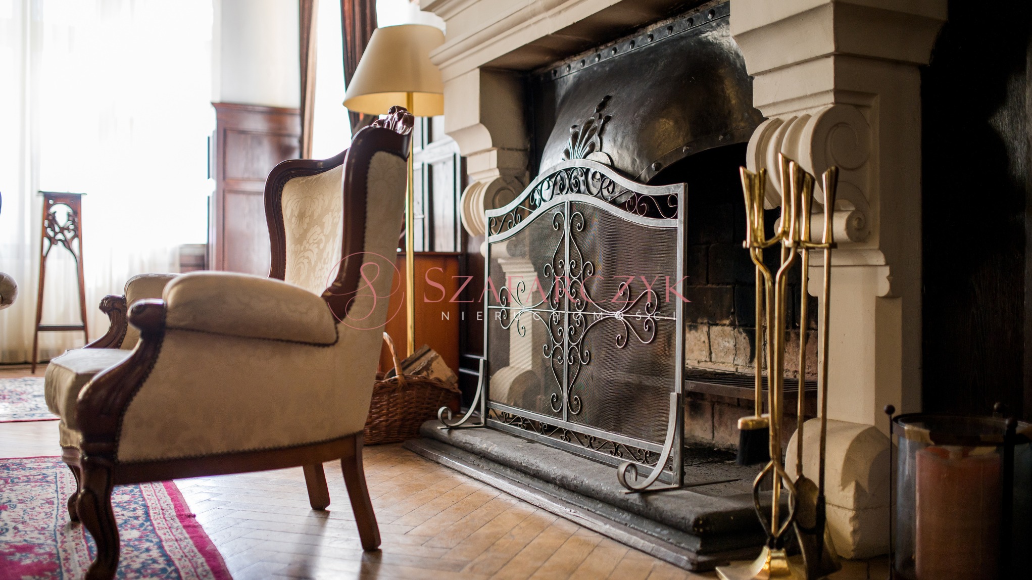 Elegancki salon z kominkiem zabezpieczonym ozdobnym, metalowym parawanem, obok którego stoją złote akcesoria kominkowe, a w tle widać stylowy fotel i lampę.