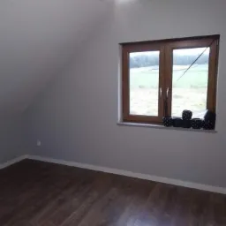 Pokój na poddaszu z drewnianą podłogą i oknem z widokiem na zielony krajobraz, ściany pomalowane na szaro, na parapecie czarne doniczki w białe kropki.