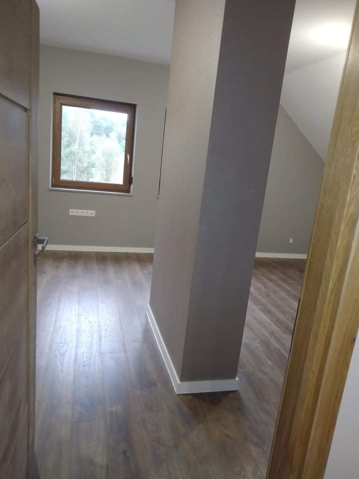 Wnętrze pokoju po remoncie: świeżo pomalowane ściany w odcieniach szarości, drewnopodobna podłoga, białe listwy przypodłogowe i drewniane okno z widokiem na drzewa.