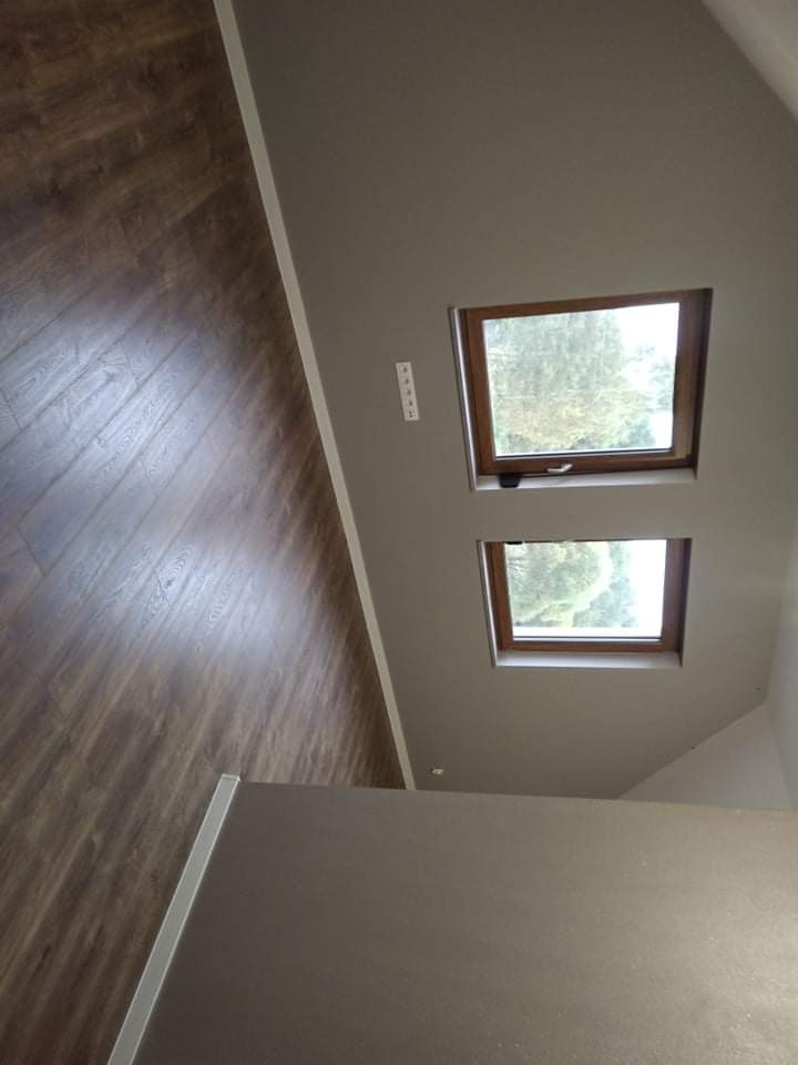 Wykończone poddasze z drewnianą podłogą, szarą ścianą i dwoma oknami dachowymi z widokiem na zieleń, widoczne gniazdka elektryczne.