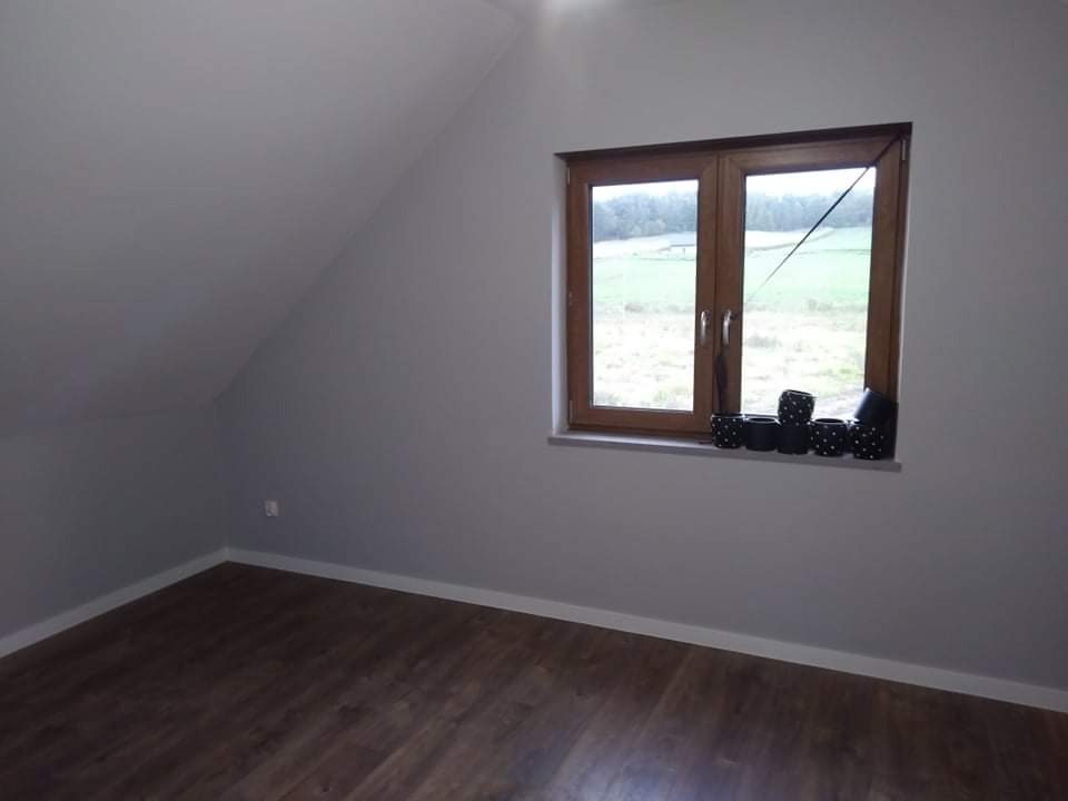 Pokój na poddaszu z drewnianą podłogą i oknem z widokiem na zielony krajobraz, ściany pomalowane na szaro, na parapecie czarne doniczki w białe kropki.