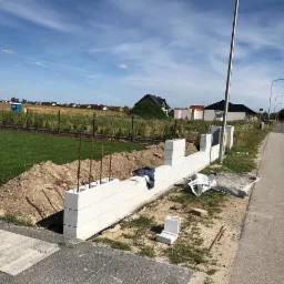 Wznoszenie muru z białych bloczków betonowych na fundamencie, widoczne wystające pręty zbrojeniowe, ziemia i ulica w tle, etap budowy ogrodzenia.