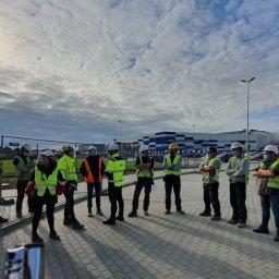 Sela Sp. z o.o. - Grupa osób w odblaskowych kamizelkach i kaskach na zewnątrz budynku, prawdopodobnie uczestnicząca w szkoleniu z zakresu bezpieczeństwa pracy na placu budowy.