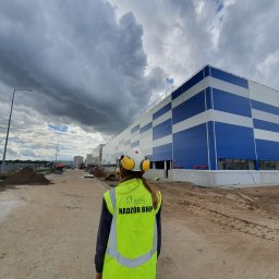 Sela Sp. z o.o. - Osoba w kamizelce odblaskowej z napisem 'Nadzór BHP' i kasku ochronnym na tle budowy hali magazynowej z niebiesko-białą elewacją w kratkę.
