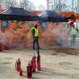 Sela Sp. z o.o. - Grupa osób w kaskach i kamizelkach odblaskowych ćwiczy gaszenie pożaru przy użyciu gaśnic, w tle namioty z grafiką płomieni i strzałką ewakuacyjną.