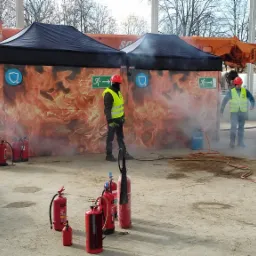 Grupa osób w kaskach i kamizelkach odblaskowych ćwiczy gaszenie pożaru przy użyciu gaśnic, w tle namioty z grafiką płomieni i strzałką ewakuacyjną.