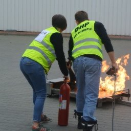 Sela Sp. z o.o. - Dwie osoby w odblaskowych kamizelkach demonstrują gaszenie płonącego ognia przy użyciu gaśnicy podczas szkolenia.