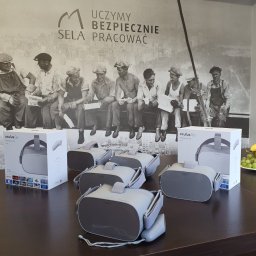 Sela Sp. z o.o. - Oculus Go VR na stole, w tle grafika z robotnikami na belce i napisem 'Uczymy bezpiecznie sela pracować', owoce na tacy z prawej strony.