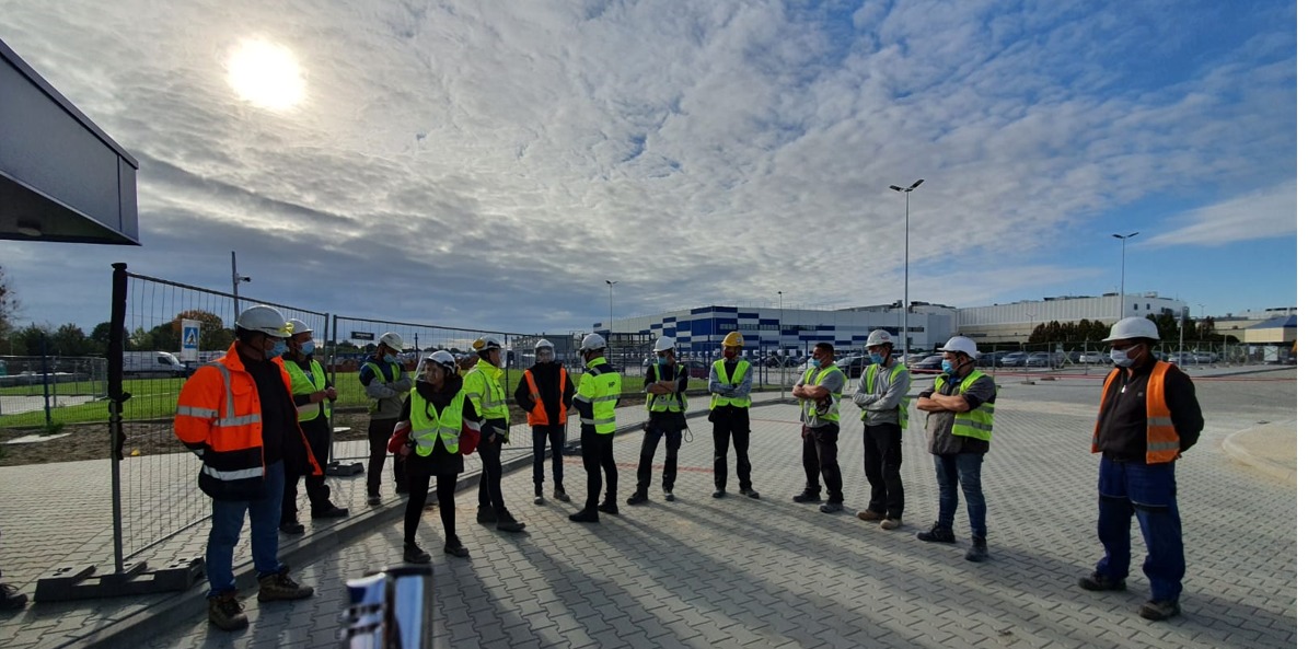 Grupa osób w odblaskowych kamizelkach i kaskach na zewnątrz budynku, prawdopodobnie uczestnicząca w szkoleniu z zakresu bezpieczeństwa pracy na placu budowy.
